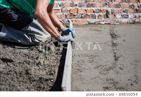 Close up of man builder placing screed rail on the floor covered with sand-cement mix at construction site on the roof. Male worker leveling surface with straight edge while screeding floor. 89850054