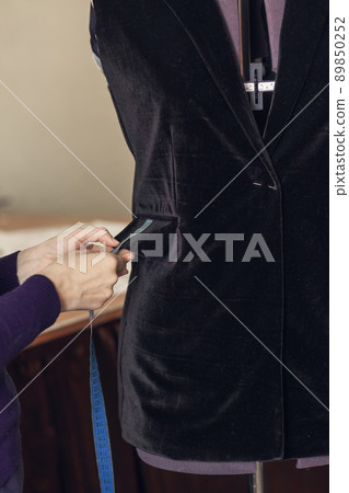 A female dressmaker sews and measures details on a black corduroy jacket hanging on a mannequin in her atelier, close-up A female dressmaker sews and measures details on a black corduroy jacket hanging on a mannequin in her atelier, close-up 89850252