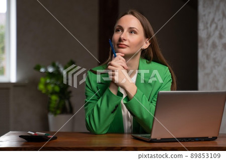Pensive woman sits at desk near laptop holding pen in hand 89853109