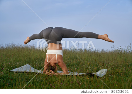 Back view of athletic strong woman standing on head on yoga mat among grass, doing exercise. Sporty female standing in asana, have static practice among nature. Concept of sport and health. 89853661