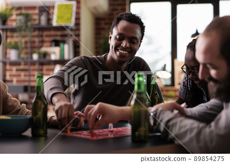 Confident smiling african american player playing boardgames with friends at home. Happy man moving plastic figurines on game table while sitting with diverse people at table. Confident smiling african american player playing boardgames with friends at home. Happy man moving plastic figurines on game table while sitting with diverse people at table. 89854275
