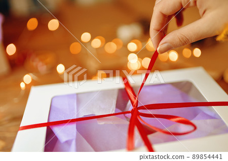 Close-up of girl's hand pulling ribbon from surprise box. Bokeh lights in background. Unpacking gift on wooden table. Festive concept of Christmas or Birthday. Close-up of girl's hand pulling ribbon from surprise box. Bokeh lights in background. Unpacking gift on wooden table. Festive concept of Christmas or Birthday. 89854441