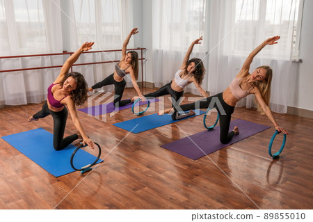 Group of young womans with magic pilates ring in Sportswear Leggings and Tops, stretching in the gym, on a yoga mat near the large window on a sunny day, female fitness yoga routine concept Group of young womans with magic pilates ring in Sportswear Leggings and Tops, stretching in the gym, on a yoga mat near the large window on a sunny day, female fitness yoga routine concept 89855010