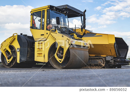 Asphalt paving machine and heavy vibratory roller close-up. A paver finisher, asphalt finisher or paving machine placing a layer of asphalt. Repaving the road surface. Road works. 89856032