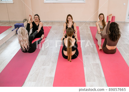 A group of six athletic women doing pilates or yoga on pink mats in front of a window in a beige loft studio interior. Teamwork, good mood and healthy lifestyle concept. 89857665