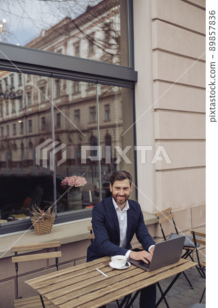 Handsome Caucasian man in elegant suit sitting at table in cafe terrace and working on laptop. Handsome Caucasian man in elegant suit sitting at table in cafe terrace and working on laptop. 89857836