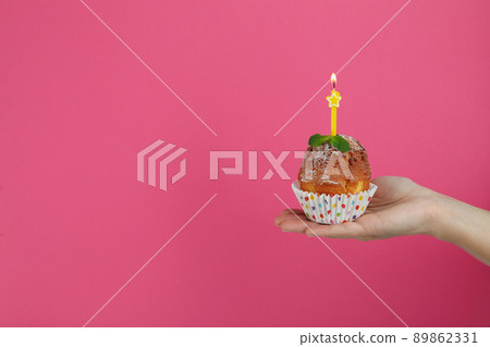 Female hand holds muffin with one candle on pink background 89862331