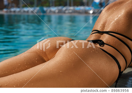 Close up of ass of slim seductive girl lying on abdomen near swimming pool. Young female wearing black swimsuit sunbathing, swimming outdoors. Concept of vacation and youth. Close up of ass of slim seductive girl lying on abdomen near swimming pool. Young female wearing black swimsuit sunbathing, swimming outdoors. Concept of vacation and youth. 89863956