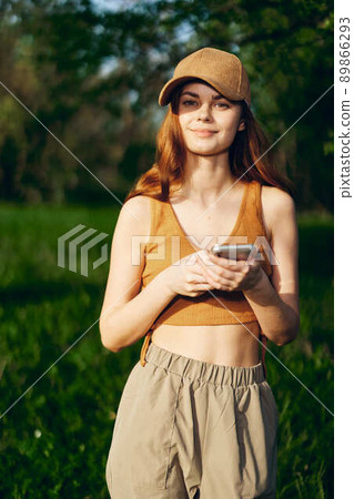 A young woman stands with her phone in her hands and smiles in the nature in the sunlight. Freelancer Lifestyle A young woman stands with her phone in her hands and smiles in the nature in the sunlight. Freelancer Lifestyle 89866293