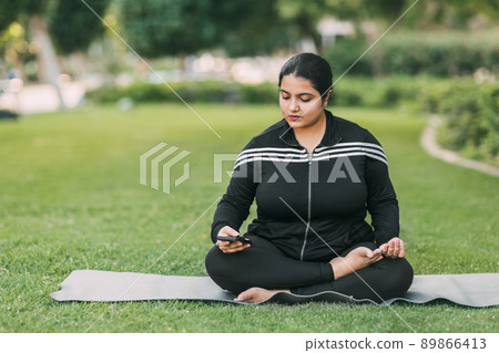 A happy young girl of Indian nationality is sitting in a lotus position in the park with a phone in her hand, resting after a yoga class 89866413