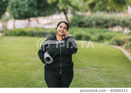 A young Indian woman with a sports mat in her hands is walking from a workout in the fresh air, talking on the phone and smiling A young Indian woman with a sports mat in her hands is walking from a workout in the fresh air, talking on the phone and smiling 89866415