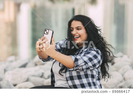 An Indian girl takes a selfie on her phone while sitting on the embankment while walking in the city. An Indian girl takes a selfie on her phone while sitting on the embankment while walking in the city. 89866417