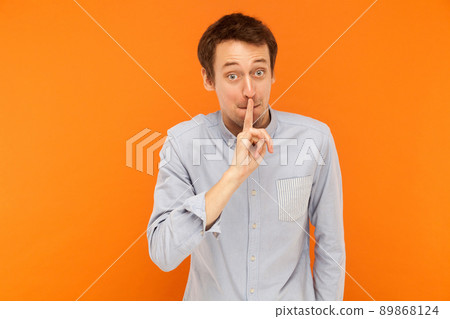 Shh, it's secret! Businessman holding finger near mouth and looking at camera with big shocked eyes. Studio shot, orange wall 89868124
