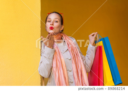 The beautiful woman with red lips, send kiss for you and holding shopping bags. Isolated studio shot on yellow background. The beautiful woman with red lips, send kiss for you and holding shopping bags. Isolated studio shot on yellow background. 89868214
