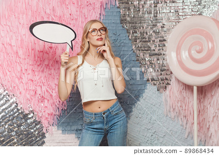 Beautiful young woman with blondie long curly hair in glasses holding speech bubble for your text and was thinking about something Beautiful young woman with blondie long curly hair in glasses holding speech bubble for your text and was thinking about something 89868434