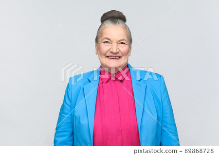 Cute woman toothy smiling at camera. Emotion and feelings Portrait of expressive grandmother with blue suit standing with collected bun gray hairstyle. Studio shot, isolated on gray background 89868728