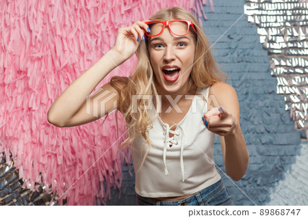 Happiness blonde girl in red glasses pointing finger and toothy smile. Indoor, studio shot, isolated on abstract background 89868747
