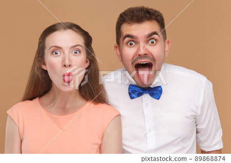 closeup portrait of funny man and beautiful woman or young couple looking at camera with amazed big eyes face and tongue out. indoor studio shot, isolated on light brown background. 89868874