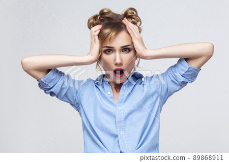 Blonde woman screaming with shock, holding hands on her head. Studio shot, gray wall 89868911