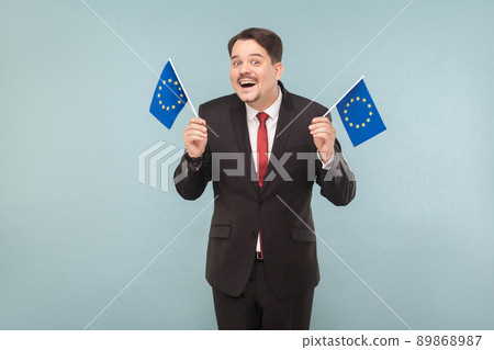 European politician holds flags and toothy smiling. Indoor, studio shot, isolated on light blue or gray background 89868987
