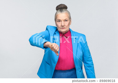 Unhappy woman showing dislike sign. Emotion and feelings, expressive grandmother with light blue suit and pink shirt standing with collected bun gray hair. Studio shot, isolated on gray background 89869018