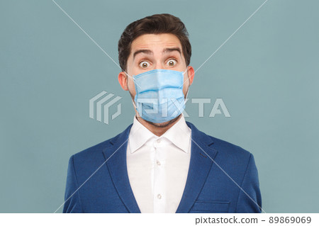 Closeup portrait of surprised or shocked young man with surgical medical mask looking at camera with big eyes. Business people medicine, health care concept. Indoor, studio shot on blue background Closeup portrait of surprised or shocked young man with surgical medical mask looking at camera with big eyes. Business people medicine, health care concept. Indoor, studio shot on blue background 89869069