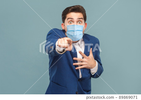 Shocked man with surgical medical mask pointing finger at camera with shocked face. Business people medicine and health care concept. Indoor, studio shot on blue background Shocked man with surgical medical mask pointing finger at camera with shocked face. Business people medicine and health care concept. Indoor, studio shot on blue background 89869071