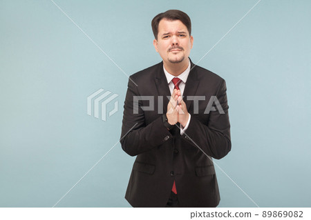 I was detained at work and asked my wife to forgive. indoor studio shot. isolated on light blue background. handsome businessman with black suit, red tie and mustache looking at camera. 89869082