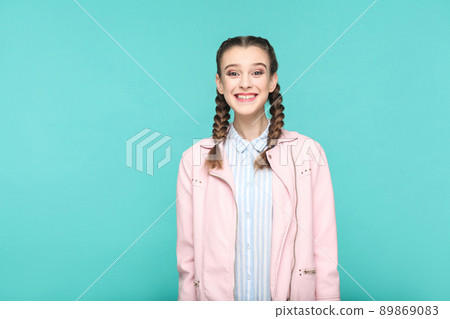happy toothy smile portrait of beautiful cute girl standing with makeup and brown pigtail hairstyle in striped light blue shirt pink jacket. indoor, studio shot isolated on blue or green background. happy toothy smile portrait of beautiful cute girl standing with makeup and brown pigtail hairstyle in striped light blue shirt pink jacket. indoor, studio shot isolated on blue or green background. 89869083