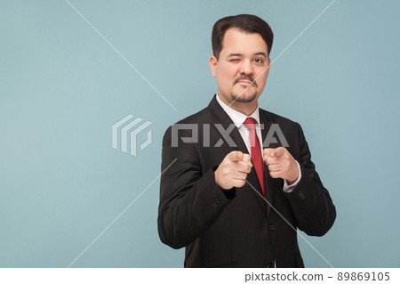 Hey you, I like you! A man winks and flirts with you. Indoor studio shot, isolated on light blue background. handsome businessman with black suit, red tie and mustache pointing and looking at camera 89869105