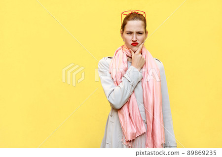 Pretty ginger girl at autumn. Wearing in coat and pink scarf, and thinking. Isolated studio shot on yellow background. Studio shot Pretty ginger girl at autumn. Wearing in coat and pink scarf, and thinking. Isolated studio shot on yellow background. Studio shot 89869235