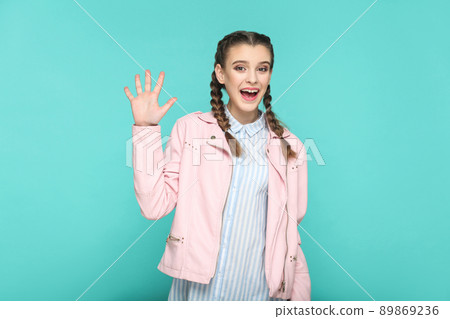 hey, amazed greeting portrait of beautiful cute girl standing with makeup and brown pigtail hairstyle in striped light blue shirt pink jacket. indoor, studio shot isolated on blue or green background. 89869236