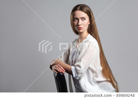 Portrait of thoughtful serious young model actor with long brown hair in white t-shirt sitting on black chair, side looking away and thinking. Indoor, studio shot. isolated on white background. 89869248