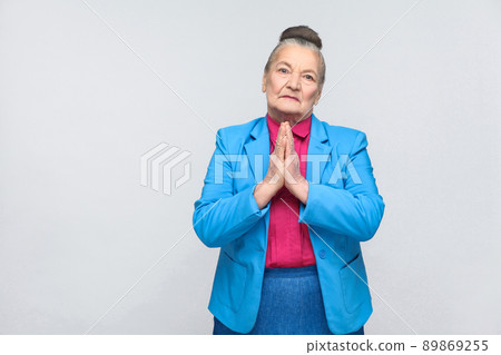 Please forgive me. Emotion and feelings concept. handsome expressive grandmother with light blue suit and pink shirt standing with collected bun gray hair. indoor, isolated on gray background Please forgive me. Emotion and feelings concept. handsome expressive grandmother with light blue suit and pink shirt standing with collected bun gray hair. indoor, isolated on gray background 89869255