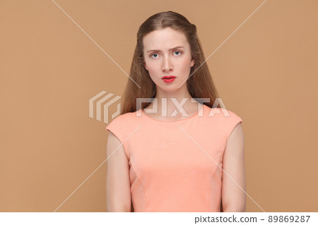 unhappy, sad woman looking at camera. portrait of emotional cute, beautiful woman with makeup and long hair in pink dress. indoor, studio shot, isolated on light brown or beige background. unhappy, sad woman looking at camera. portrait of emotional cute, beautiful woman with makeup and long hair in pink dress. indoor, studio shot, isolated on light brown or beige background. 89869287