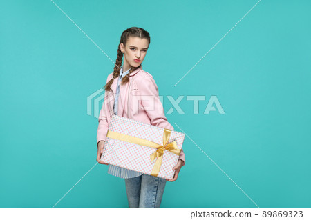 Funny beautiful girl in casual style, pigtail hairstyle and pink jacket, standing and holding dotted gift box and looking at camera with kiss, Indoor, studio shot isolated on blue or green background 89869323