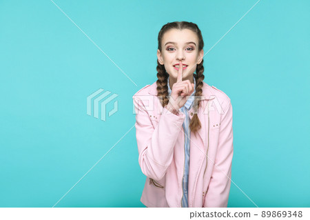 secret finger, portrait of beautiful cute girl standing with makeup and brown pigtail hairstyle in striped light blue shirt pink jacket. indoor, studio shot isolated on blue or green background. 89869348