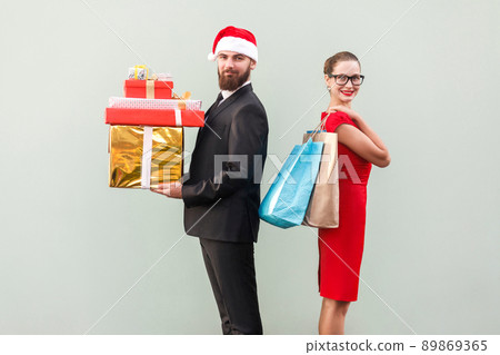 Back to back. Profile view man in red hat and woman in red dress and glasses. Happiness couple holding christmas gifts and colorful packages and looking at camera with toothy smiling. Studio shot Back to back. Profile view man in red hat and woman in red dress and glasses. Happiness couple holding christmas gifts and colorful packages and looking at camera with toothy smiling. Studio shot 89869365