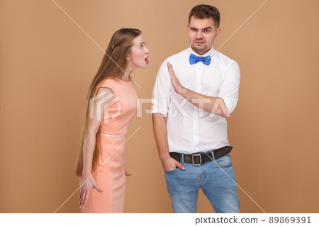 Oh please stop. confused man in white shirt try to ban, block or stop angry woman in pink dress who nervous, yelling screaming at him. indoor studio shot, isolated on light brown background. 89869391