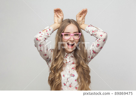Comic woman in pink glasses holding hands look's like rabbit ears, toothy smiling and looking at camera. Studio shot, gray background 89869475