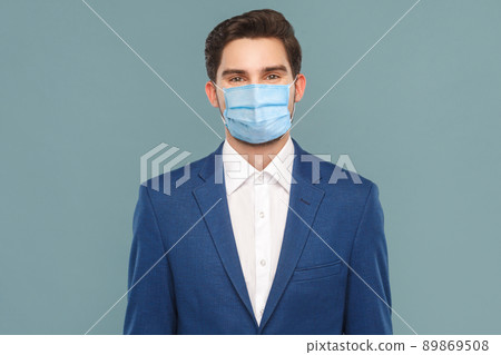 Portrait of handsome young businessman with surgical medical mask looking at camera. Business people medicine and health care concept. Indoor, studio shot on blue background Portrait of handsome young businessman with surgical medical mask looking at camera. Business people medicine and health care concept. Indoor, studio shot on blue background 89869508