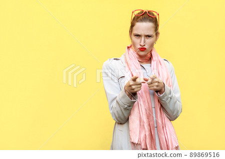 The serious woman, wearing coat, having anger looks, pointing finger at camera. Isolated shot on yellow wall The serious woman, wearing coat, having anger looks, pointing finger at camera. Isolated shot on yellow wall 89869516