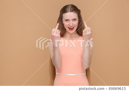 crazy woman shows middle fingers and looking at camera. portrait of emotional cute, beautiful woman with makeup and long hair in pink dress, studio shot, isolated on light brown or beige background. 89869615