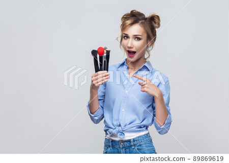 Beautiful blonde girl, holding and pointing finger to many pofessional brush, looking at camera. Isolated on gray background Beautiful blonde girl, holding and pointing finger to many pofessional brush, looking at camera. Isolated on gray background 89869619