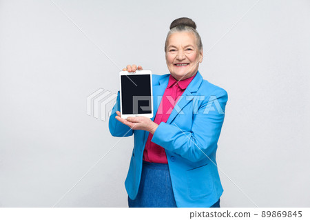 Aged woman holding and showing tablet screen with toothy smiling and satisfied face. Grandmother in light blue suit with collected gray hair bun hairstyle. Studio shot, isolated on gray background Aged woman holding and showing tablet screen with toothy smiling and satisfied face. Grandmother in light blue suit with collected gray hair bun hairstyle. Studio shot, isolated on gray background 89869845