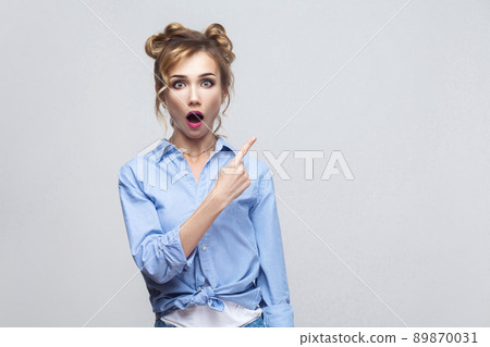 Excited blonde woman pointing finger sideways, raising eyebrows and keeping mouth wide opened, showing something surprising on grey copy space wall. Indoor, studio shot Excited blonde woman pointing finger sideways, raising eyebrows and keeping mouth wide opened, showing something surprising on grey copy space wall. Indoor, studio shot 89870031