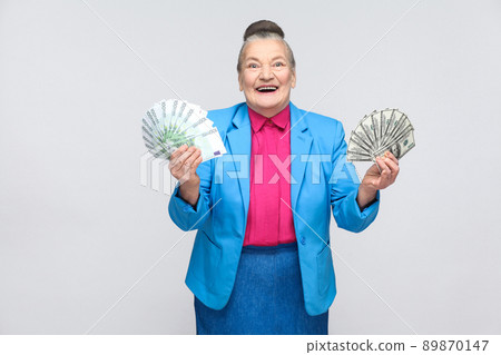 happy stylish aged woman holding many euro and dollars. Portrait of handsome expressive grandmother in light blue suit with collected gray hair bun hairstyle. Studio shot, isolated on gray background 89870147
