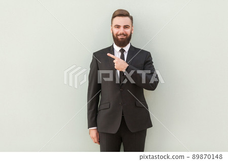Young handsome bearded businessman with beaming smile pointing with finger at capy space and looking at camera. Indoor shot, gray background 89870148
