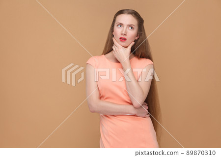confused, thoughtful woman looking away. portrait of emotional cute, beautiful woman with makeup and long hair in pink dress. indoor, studio shot, isolated on light brown or beige background. confused, thoughtful woman looking away. portrait of emotional cute, beautiful woman with makeup and long hair in pink dress. indoor, studio shot, isolated on light brown or beige background. 89870310