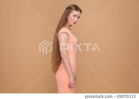 unhappy sad woman with long brown hair looking at camera. emotional cute, beautiful woman with makeup and long hair in pink dress. indoor, studio shot, isolated on light brown or beige background. 89870315